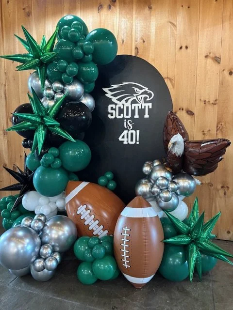Balloon display with footballs, green and silver balloons, and a black sign that says "Scott is 40!" with a lion logo, set against a wooden wall.
