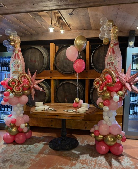 Table with pink, gold, and white balloons, including large star-shaped balloons and cake-shaped balloons with "20" and "Say Pop" written on them, set in front of wine barrels for a celebration.