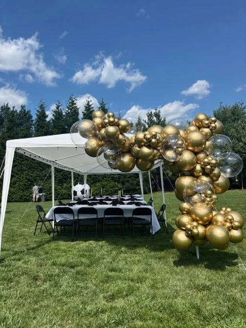Outdoor party setup with a white canopy tent, covered tables, black chairs, and a balloon arch of gold and transparent balloons, with a grassy field and trees in the background under a blue sky.