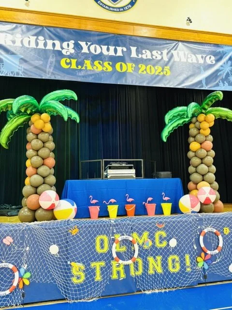 Decorated stage with inflatable palm trees, beach balls, and a banner that reads 'Riding Your Last Wave, Class of 2025'. The front of the stage has a blue net with the words 'OLMC STRONG!' in yellow. There are colorful cups and flamingo decorations o