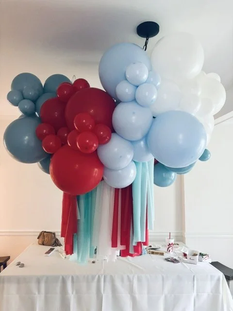 Cluster of multicolored balloons including blue, white, and red, with hanging ribbons beneath, indoors over a table.