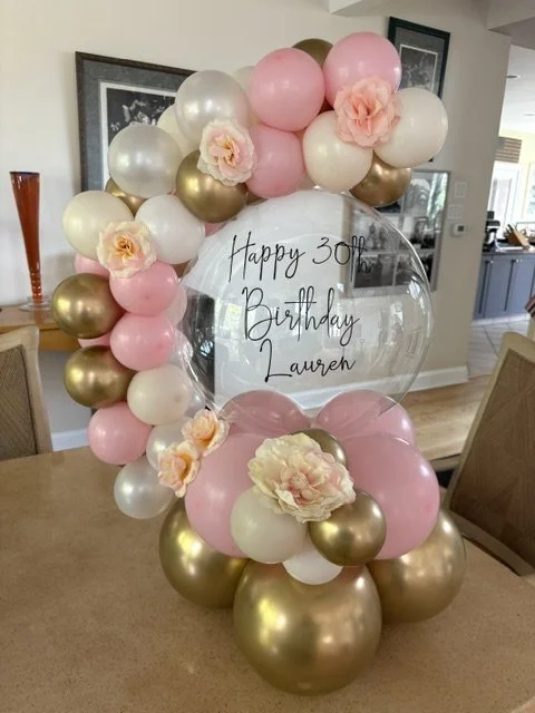 Balloon arrangement with pink, white, and gold balloons, pink flowers, and a clear balloon with 'Happy 30th Birthday Lauren' written inside, set on a table in a dining area.