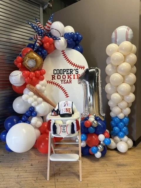 Baseball-themed birthday decorations with balloons in red, blue, and white, featuring a large baseball graphic with the text 'Cooper's Rookie Year' and a silver number one balloon, a baseball cap, and a baseball bat prop.