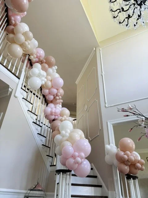 Decorative pink, white, and nude balloons arranged along a staircase railing and on top of pedestals in a well-lit interior space provides the sweetest entryway to a graduation party!