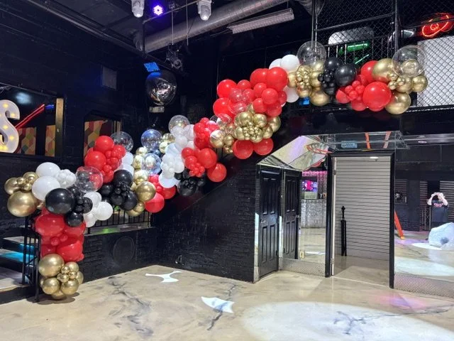A balloon arch decoration with red, white, black, gold, and transparent balloons at the entrance of a party or event space.