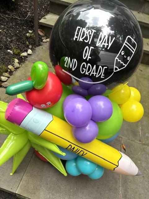 Colorful balloon arrangement with a large black balloon reading "First Day of 2nd Grade," an inflatable apple, a yellow crayon, and purple, green, and yellow balloons, celebrating a child's first day of second grade.