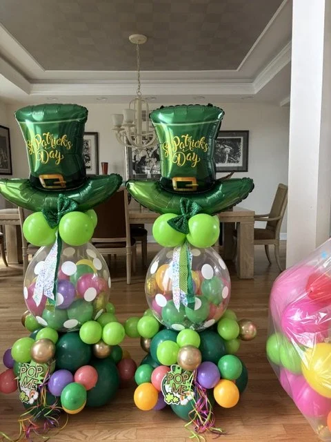 Two St. Patrick's Day balloon arrangements with green leprechaun hats on top, surrounded by colorful balloons, in a dining room.