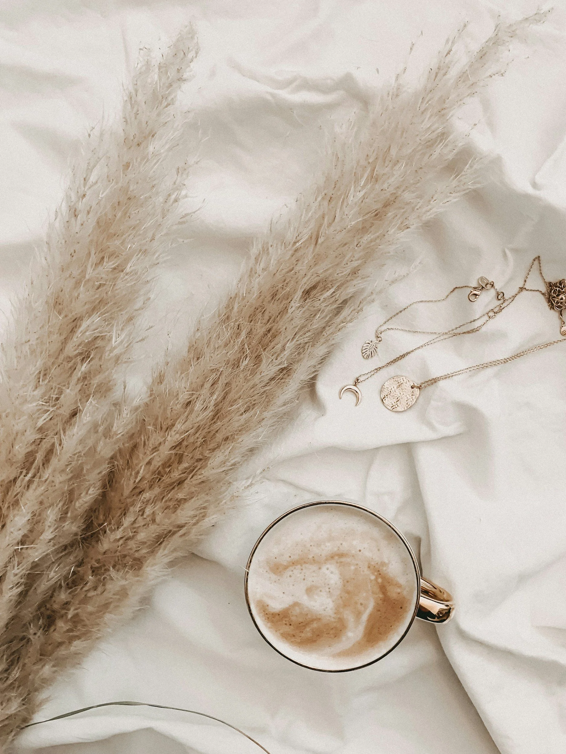 Close-up of a cup of frothy coffee, dried pampas grass, and layered necklaces with moon, leaf, and round pendant charms on a white fabric surface.