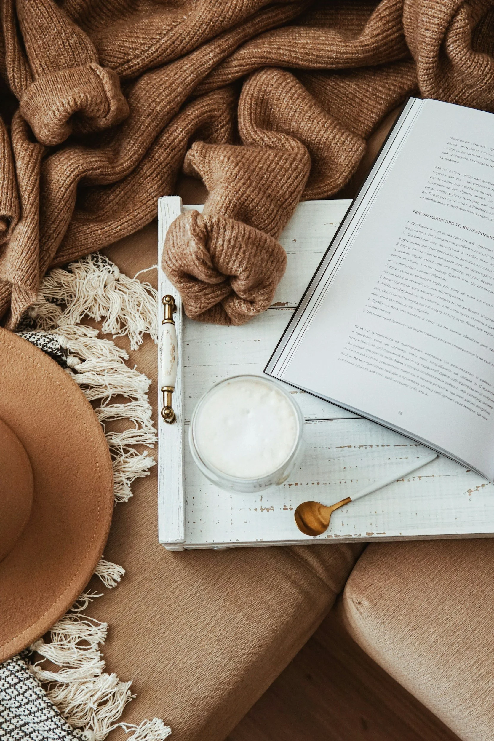 Cozy flat lay with beige hat, fringed scarf, brown knit blanket, book, glass of frothy drink, and gold spoon on white wooden tray, surrounded by beige and black-and-white textured pillows and a tan sofa.