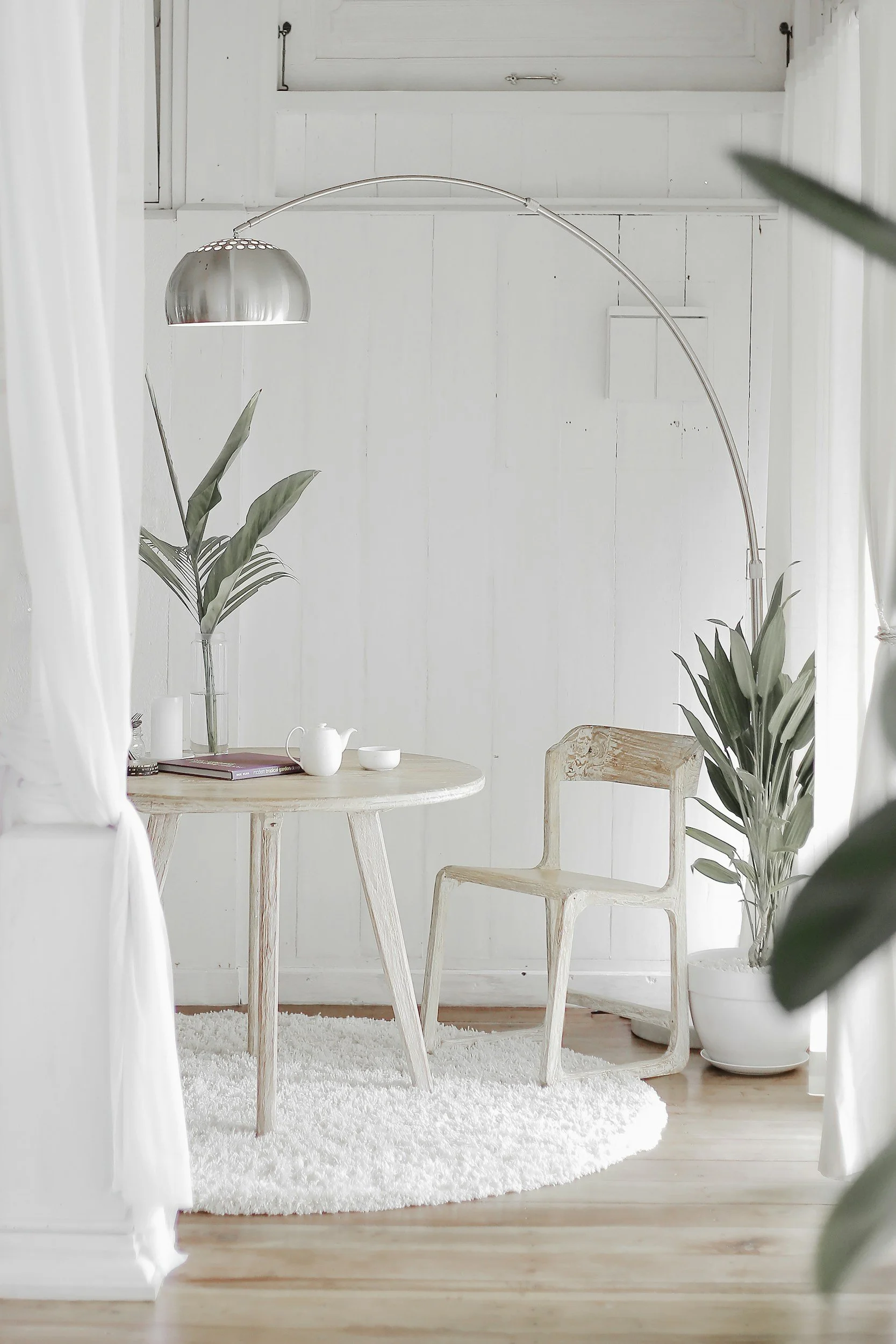 A bright, minimalistic dining area with white walls, wooden table, and chairs. Decorated with potted plants, white curtains, and a white fluffy rug on wooden flooring.