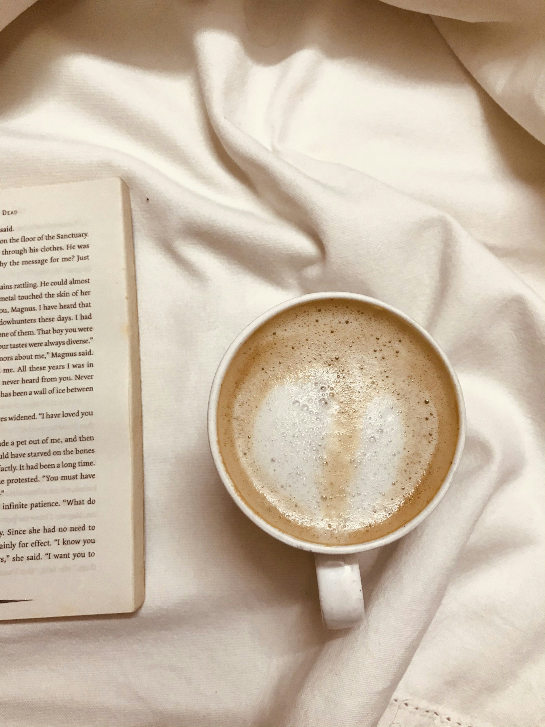 A white ceramic mug filled with coffee and foam, positioned on a cream-colored fabric surface. Part of an open book with printed text is visible on the left side of the image.