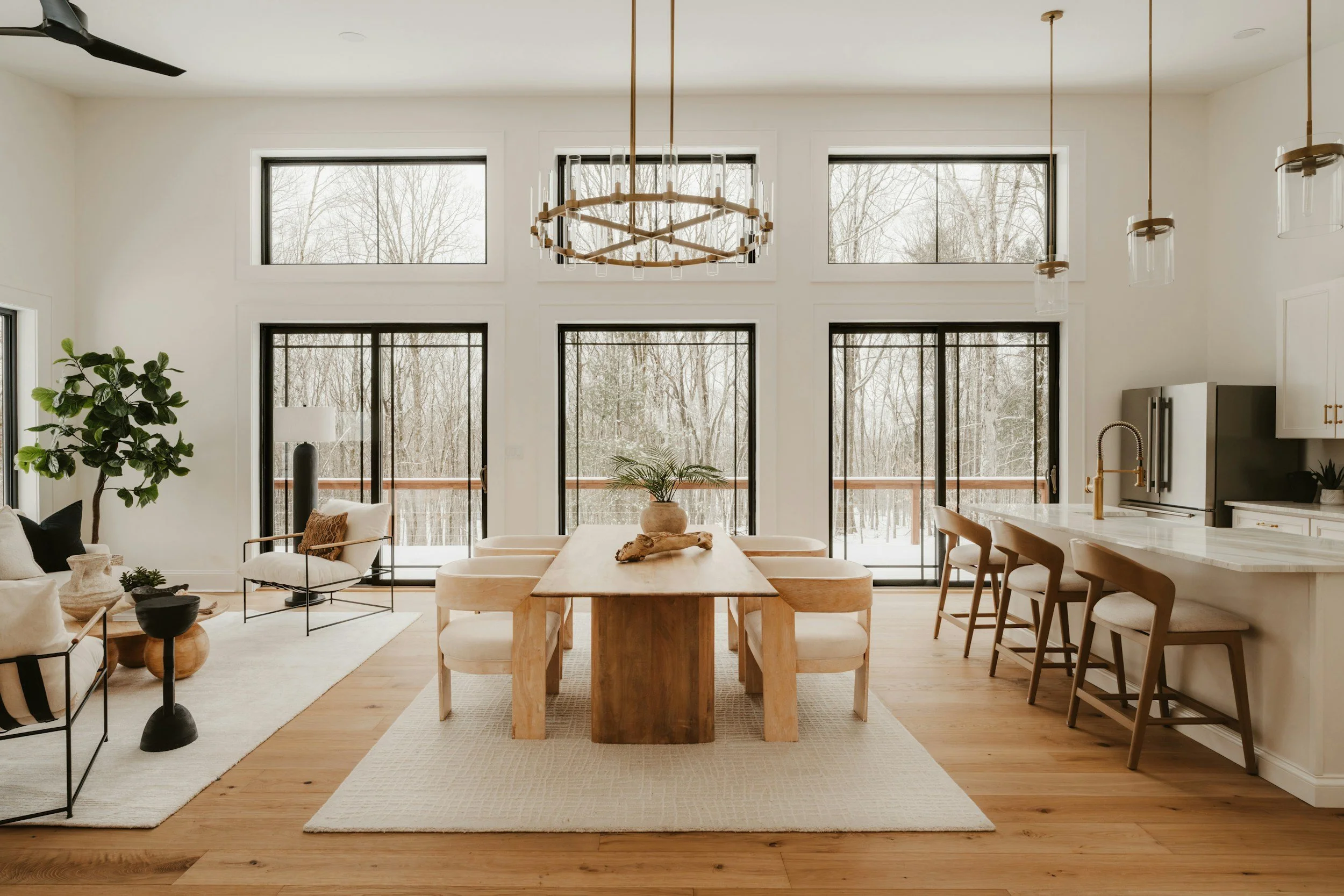 A modern, open-concept living and dining area with large windows overlooking a wintry outdoor landscape. The space features a wooden dining table with cream-colored chairs, a white sofa, a black armchair, and various decorative elements including a large potted plant, vases, and a chandelier. The kitchen area is visible with white cabinets, a black refrigerator, and a gold faucet.