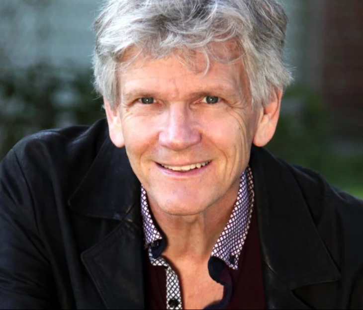 A smiling middle-aged man with gray hair, wearing a black jacket over a checkered shirt, outdoors with a blurred background.