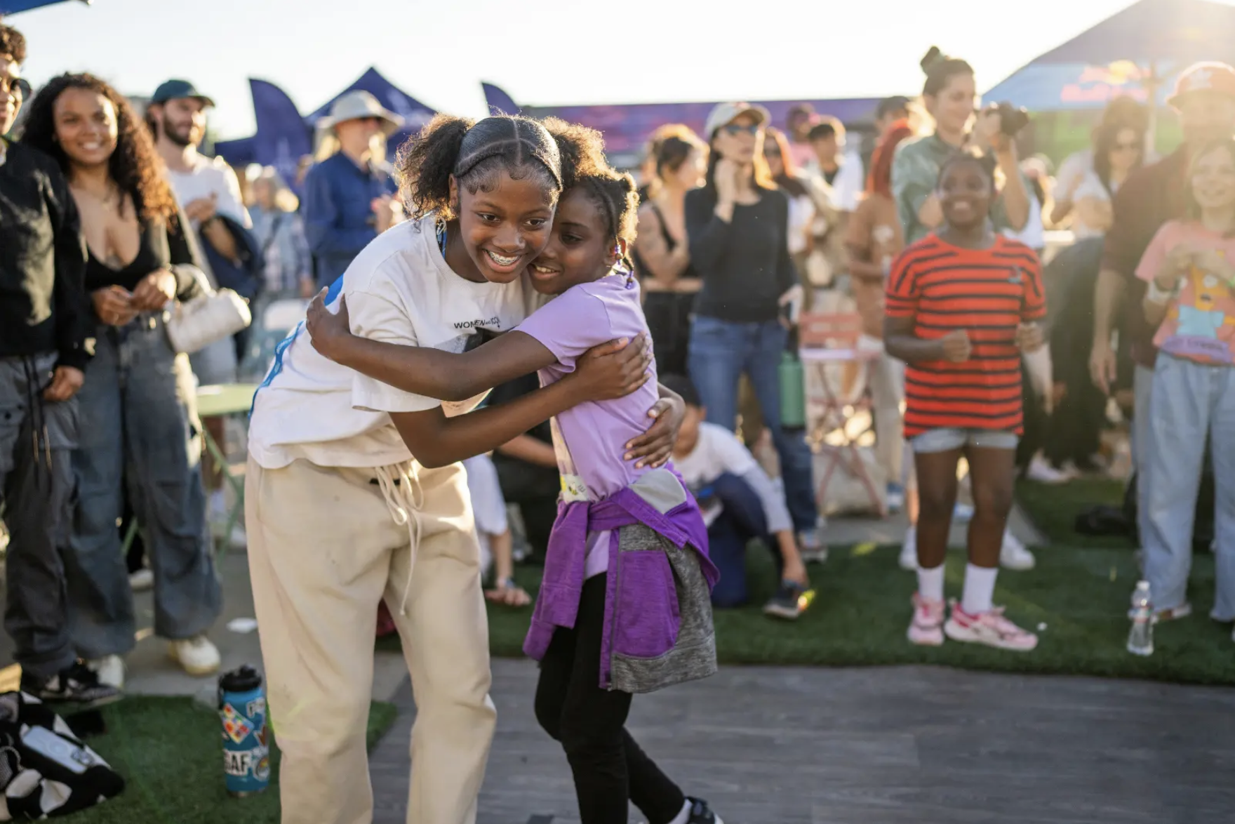 Two young girls hugging and smiling at an outdoor event with a crowd of people in the background.