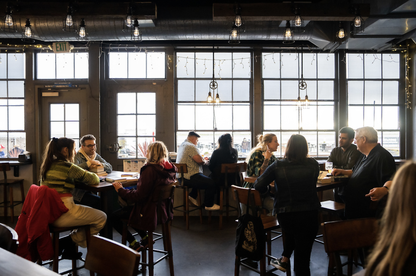 Group of people sitting inside a cafe or restaurant near large windows, engaged in conversation and reading.