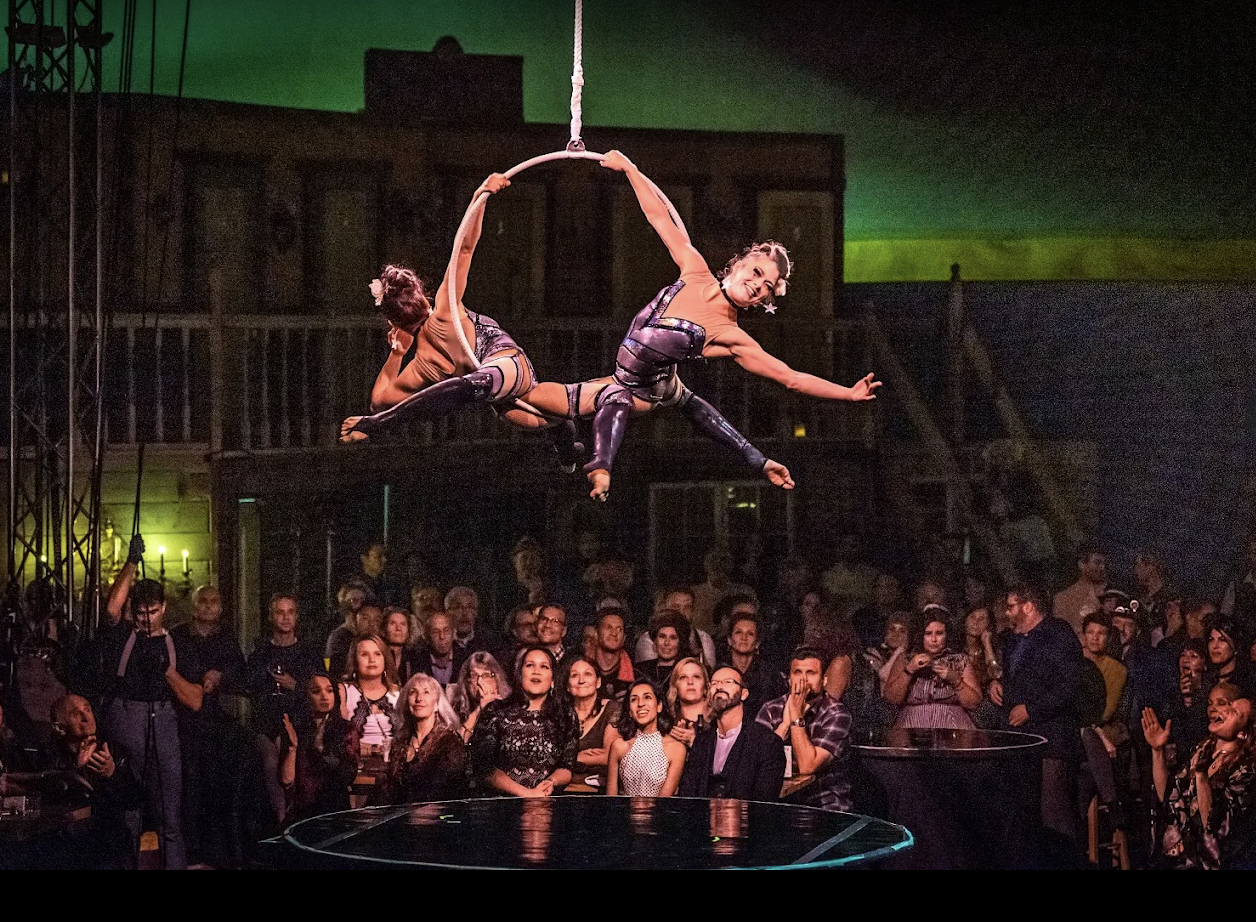 Two female aerial performers in metallic costumes perform an acrobatic routine on a hoop suspended in the air, while an audience watches them from below in a dimly lit theater.