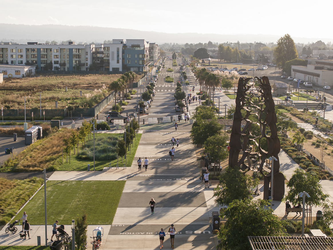 Aerial view of a wide pedestrian walkway with people walking and jogging, lined with trees and sculptures, with residential buildings, parking lots, and distant mountains in the background.