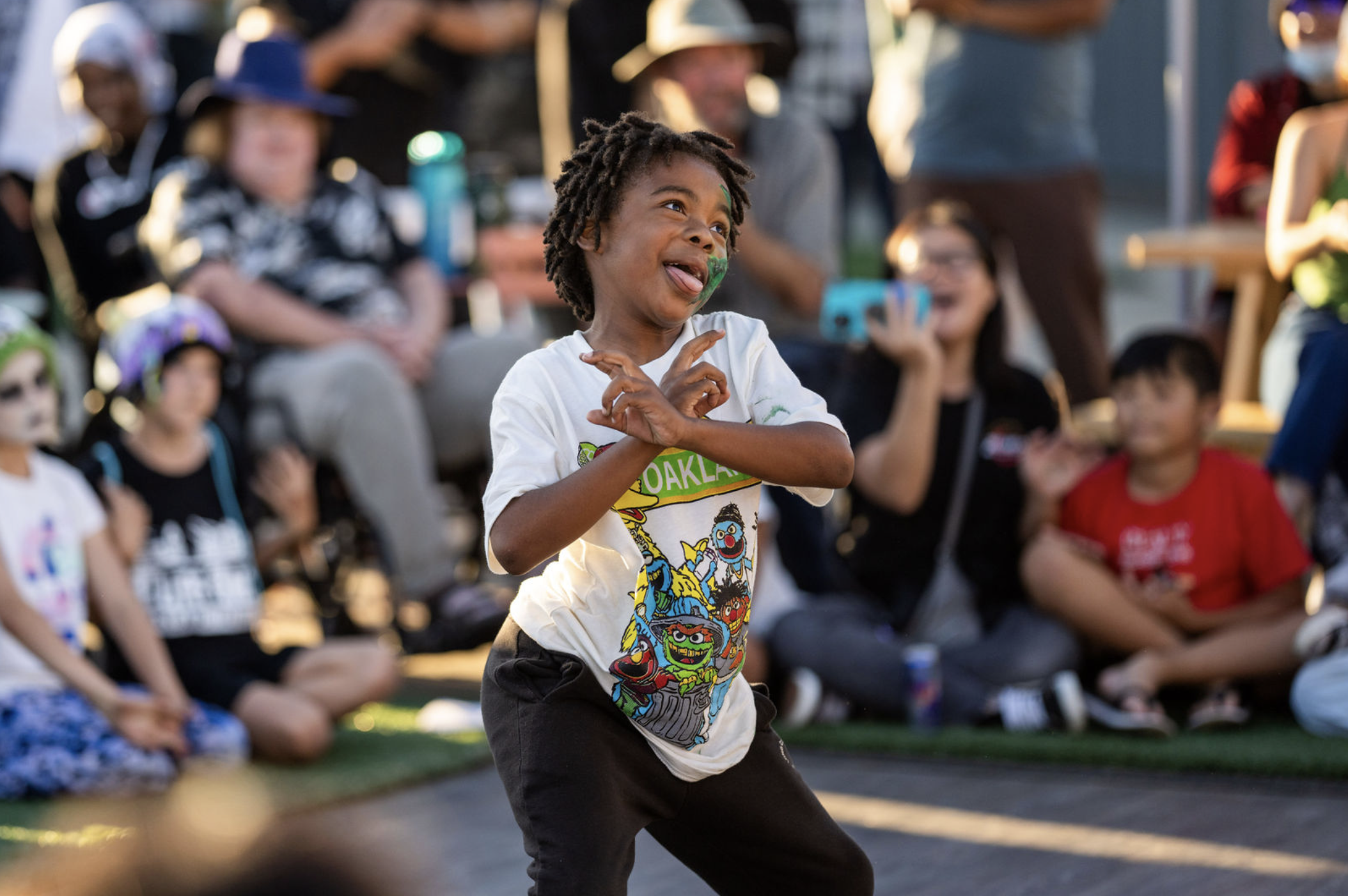 A young boy with face paint, dancing and smiling at an outdoor gathering with seated children and adults in the background.