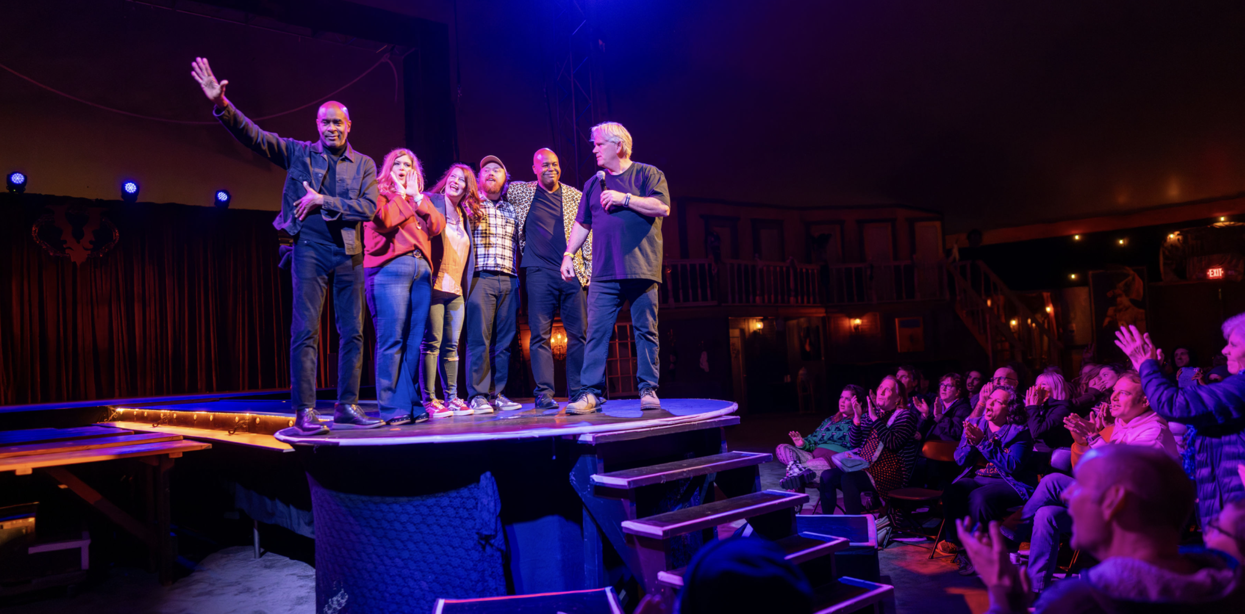 Group of performers on stage waving to audience in a comedy show, with audience clapping and smiling.