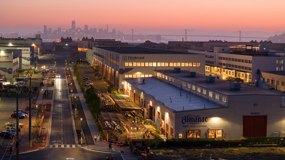 Cityscape view of a modern industrial area at dusk, with buildings, parking lots, and a distant skyline including the Transamerica Pyramid and a suspension bridge. The foreground shows a brewery with outdoor seating.