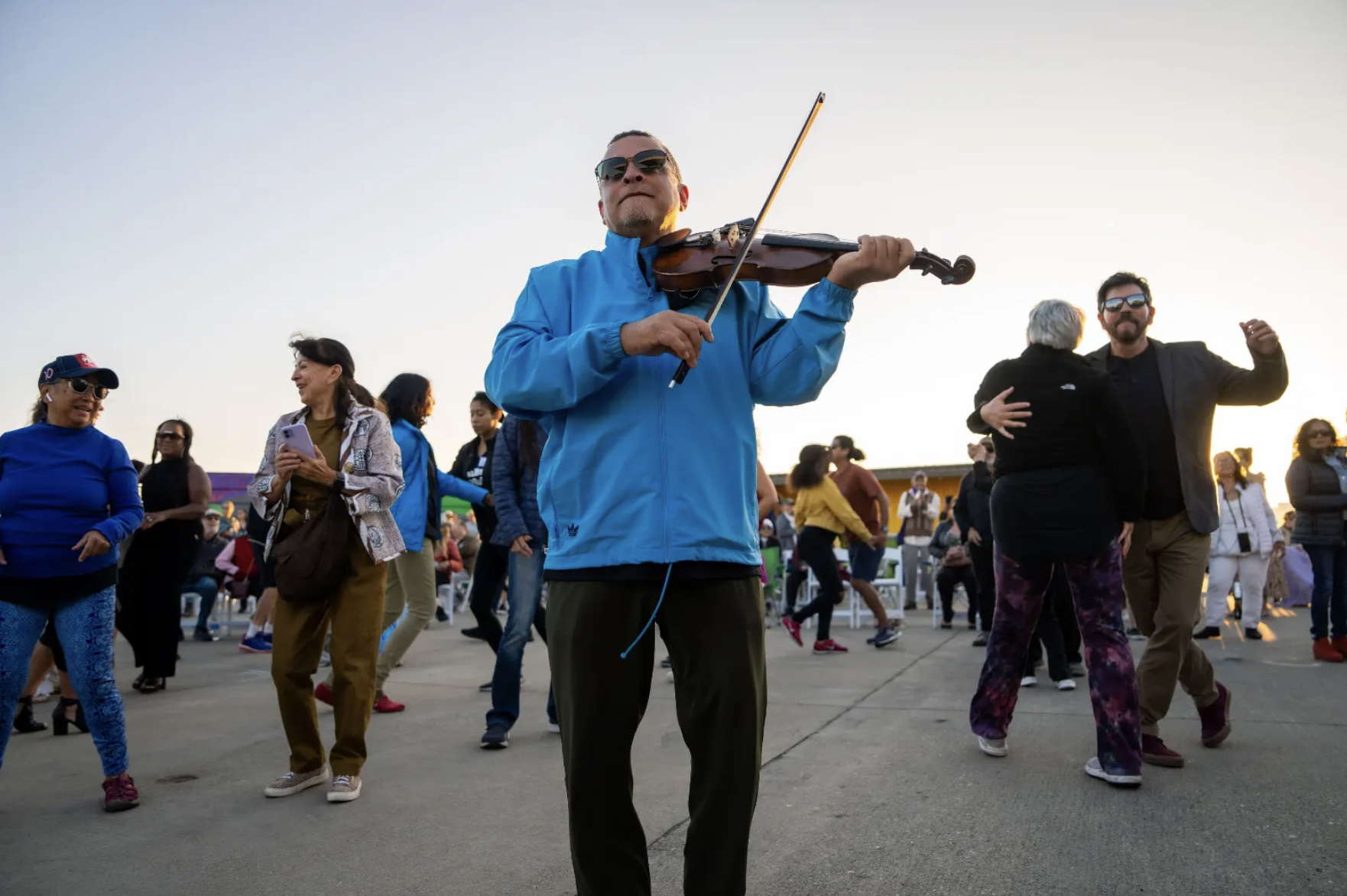 A man wearing a blue jacket and sunglasses playing a violin among a crowd of people dancing outdoors during sunset.