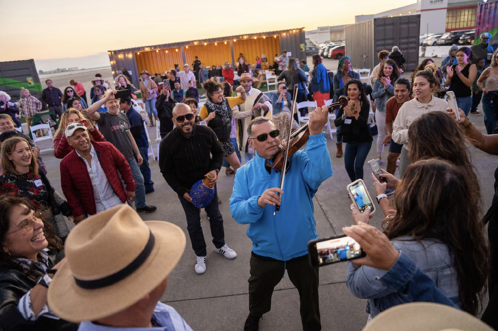 A lively outdoor gathering with people dancing and singing, led by a man playing a violin, with others taking photos and enjoying the event in the evening.
