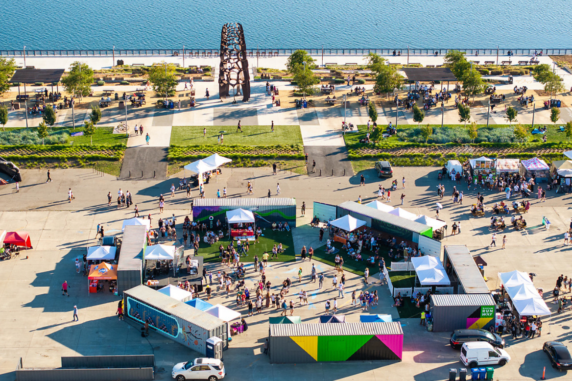 Aerial view of an outdoor event with booths, tents, and people, near a waterfront with a paved walkway and benches.