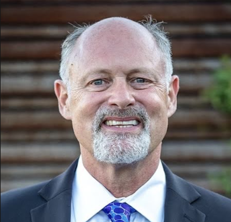A smiling man with a beard and light hair, wearing a suit and tie, outdoors in front of a wooden fence.