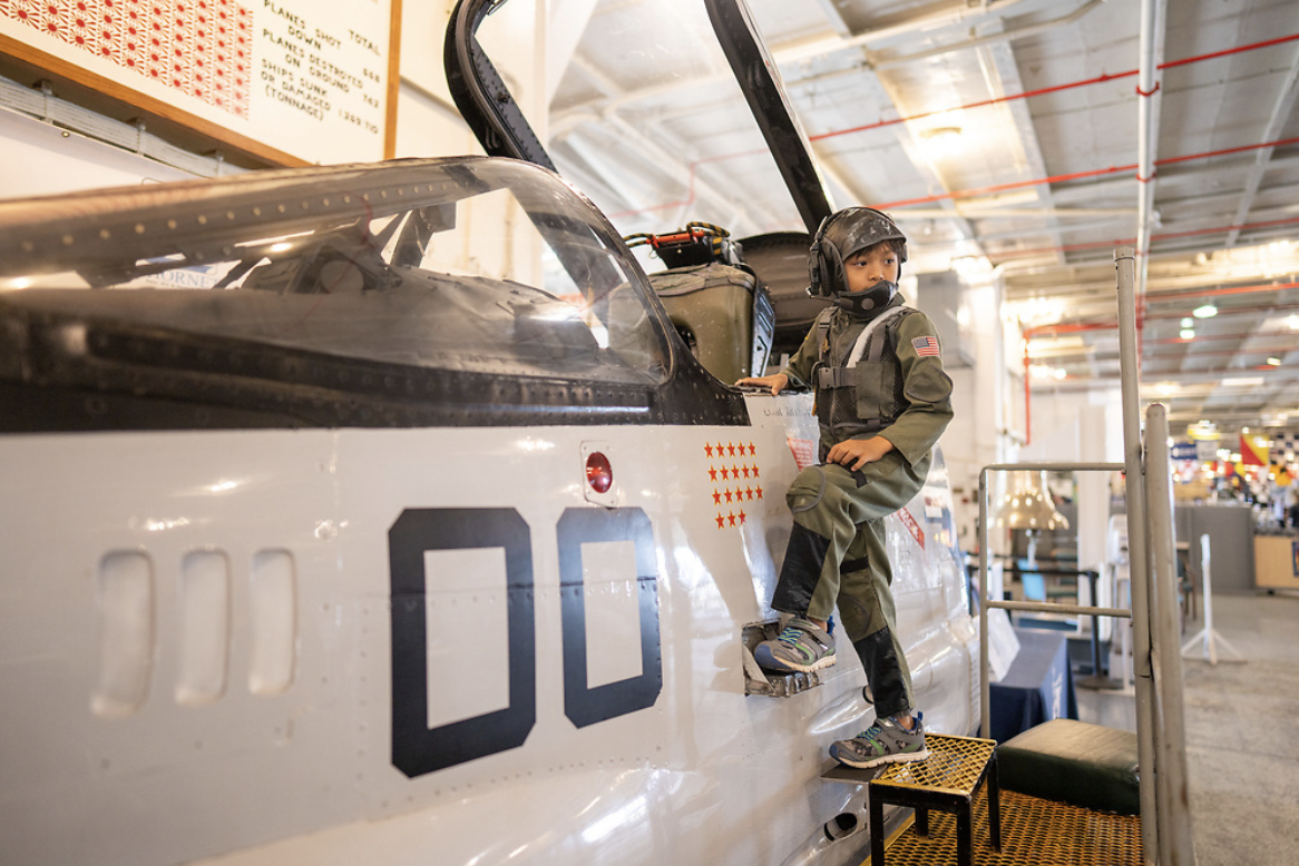 A young boy dressed in a pilot uniform with a helmet and mask, sitting on the side of a fighter jet inside a museum or exhibit.