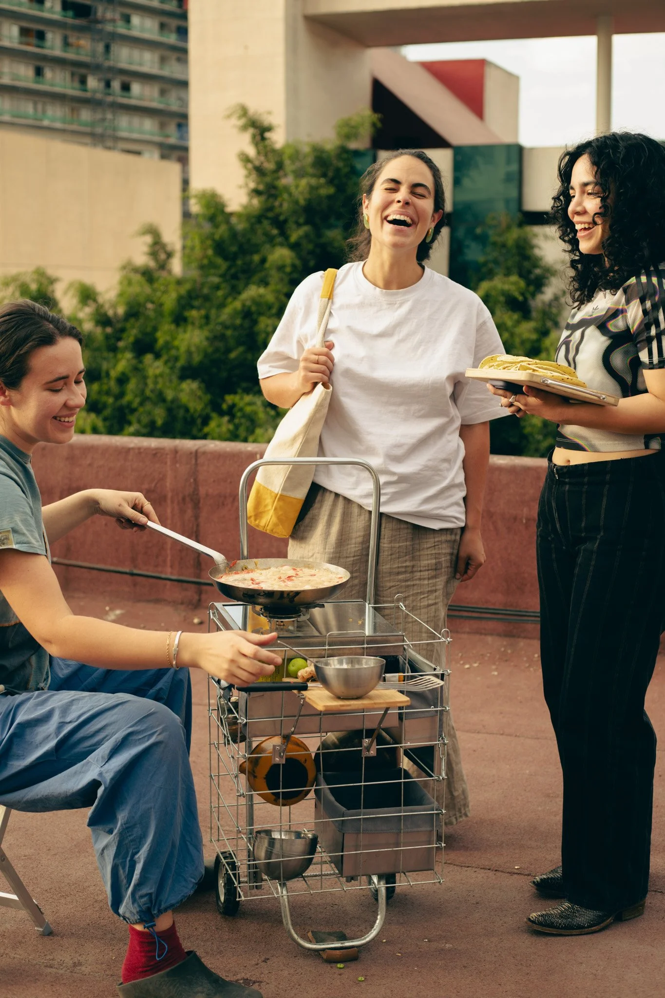 Four women enjoying outdoor food activity with a portable grill, some are sitting while others stand including a server holding a plate of hot dogs.
