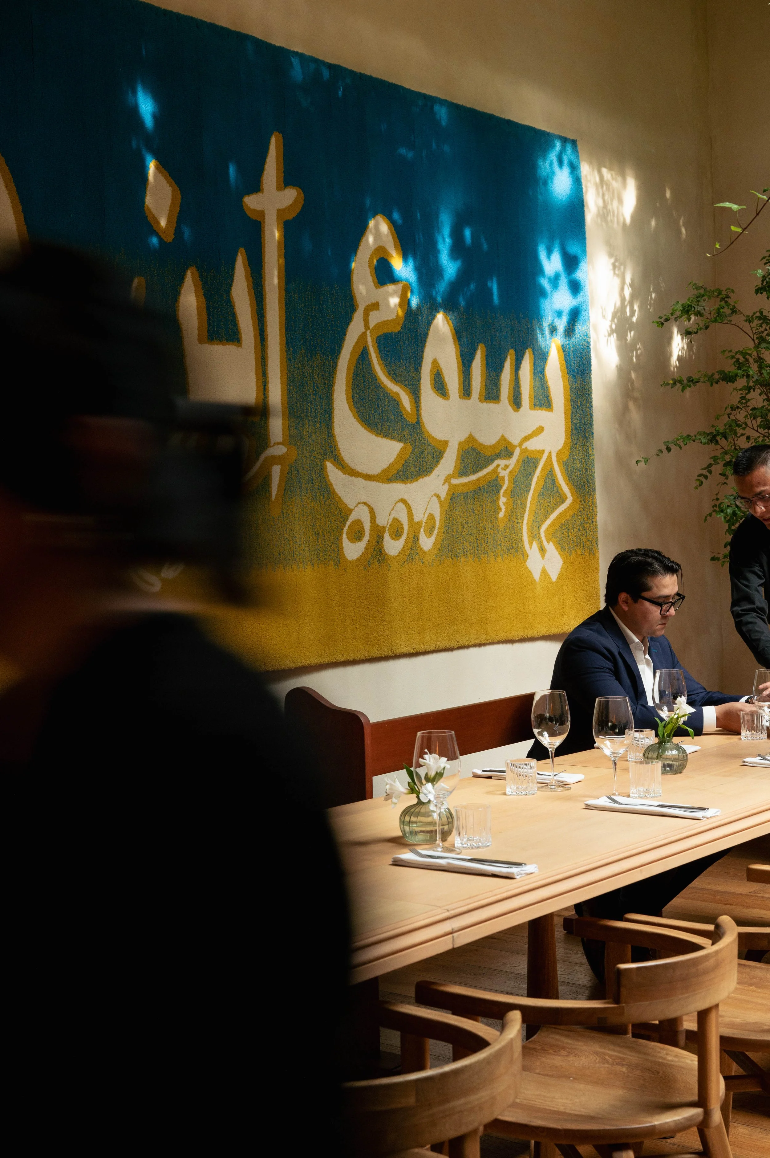 A restaurant scene with a large wall hanging featuring a stylized illustration of a donkey with a sword. People are seated at a long wooden table with wine glasses, white napkins, and small floral arrangements. There are two men visible, one sitting and the other standing, engaged in conversation.