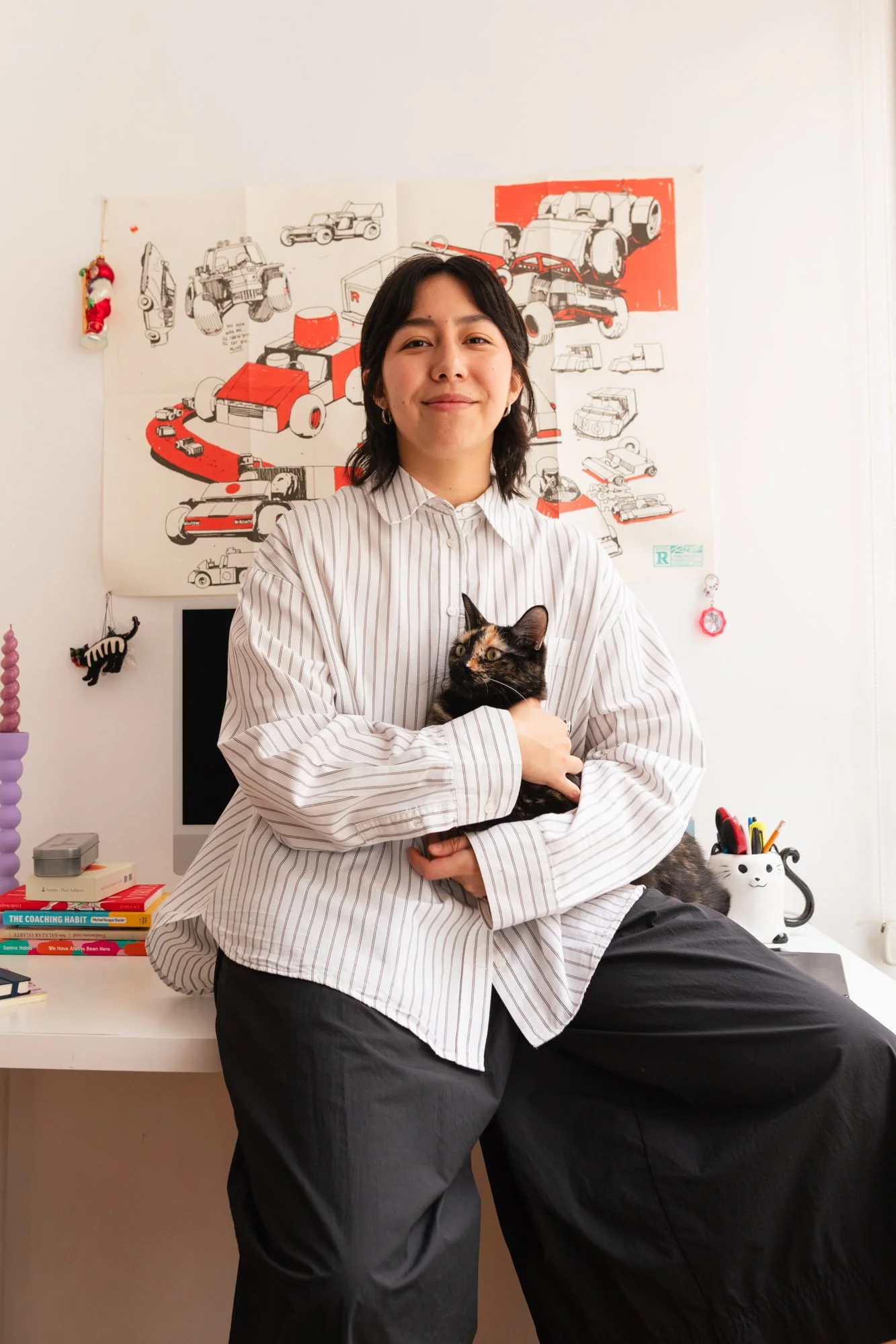 A woman in a striped shirt sitting on a desk, holding a calico cat. Behind her is a poster with sketches of cars, a desktop computer, and a collection of books and decorations in a room with white walls.