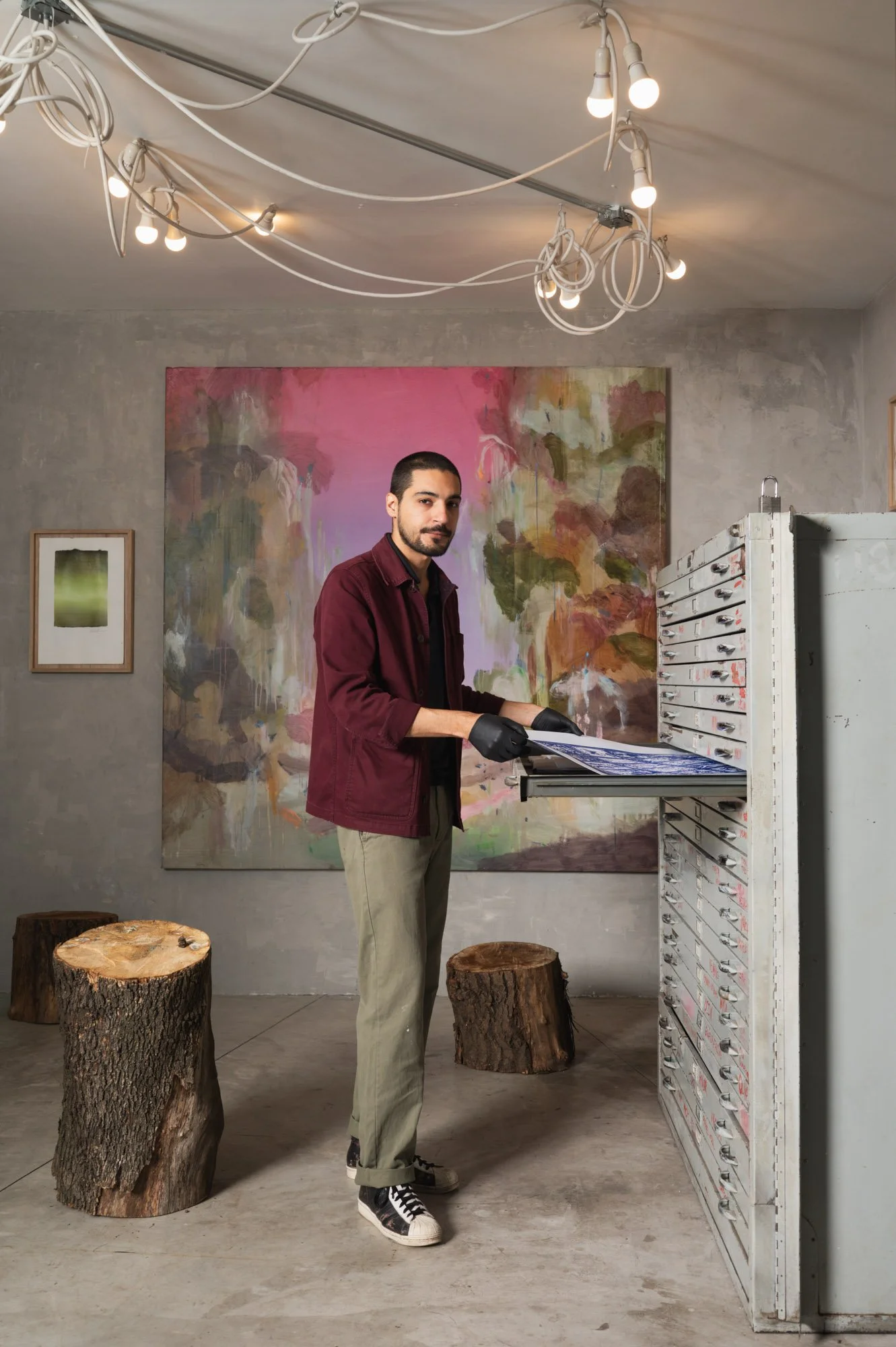A man standing in art gallery beside a white multi-drawer cabinet, wearing black gloves, burgundy shirt, beige pants, black and white sneakers, with a large colorful abstract painting behind him. There are three tree stump stools in front of him and a ceiling light fixture with exposed bulbs above.