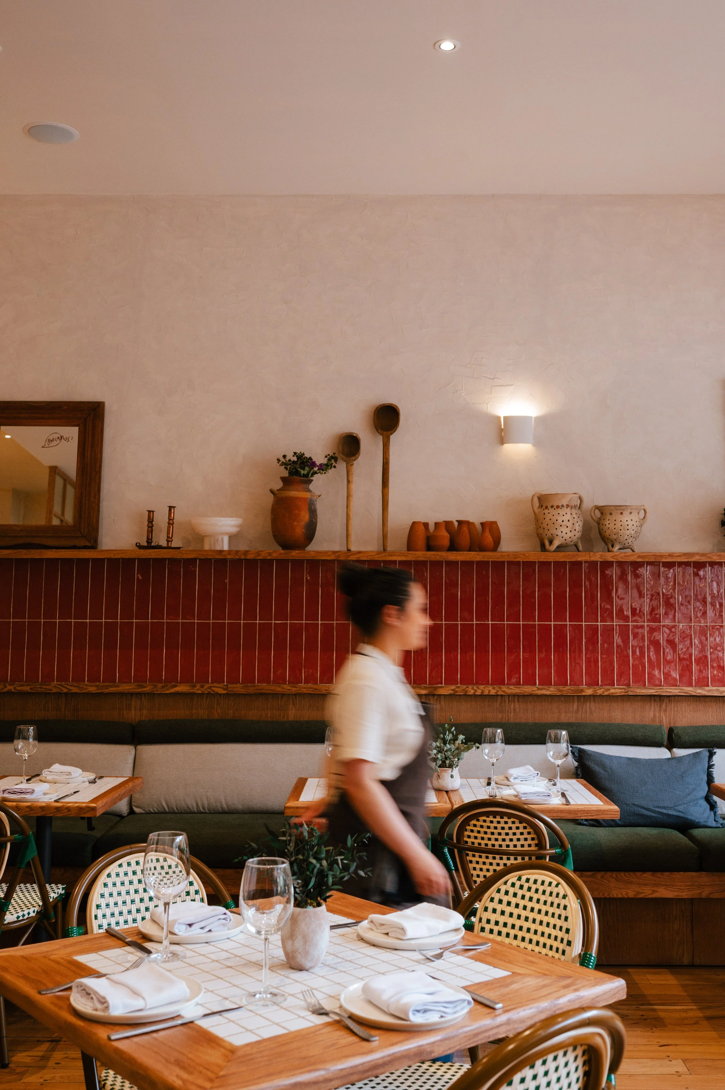 Interior of a restaurant with a table set for dining, including wine glasses, plates, napkins, and cutlery, and a woman walking past. The wall has decorative pottery and wooden utensils, with soft lighting and a mirror in the background.