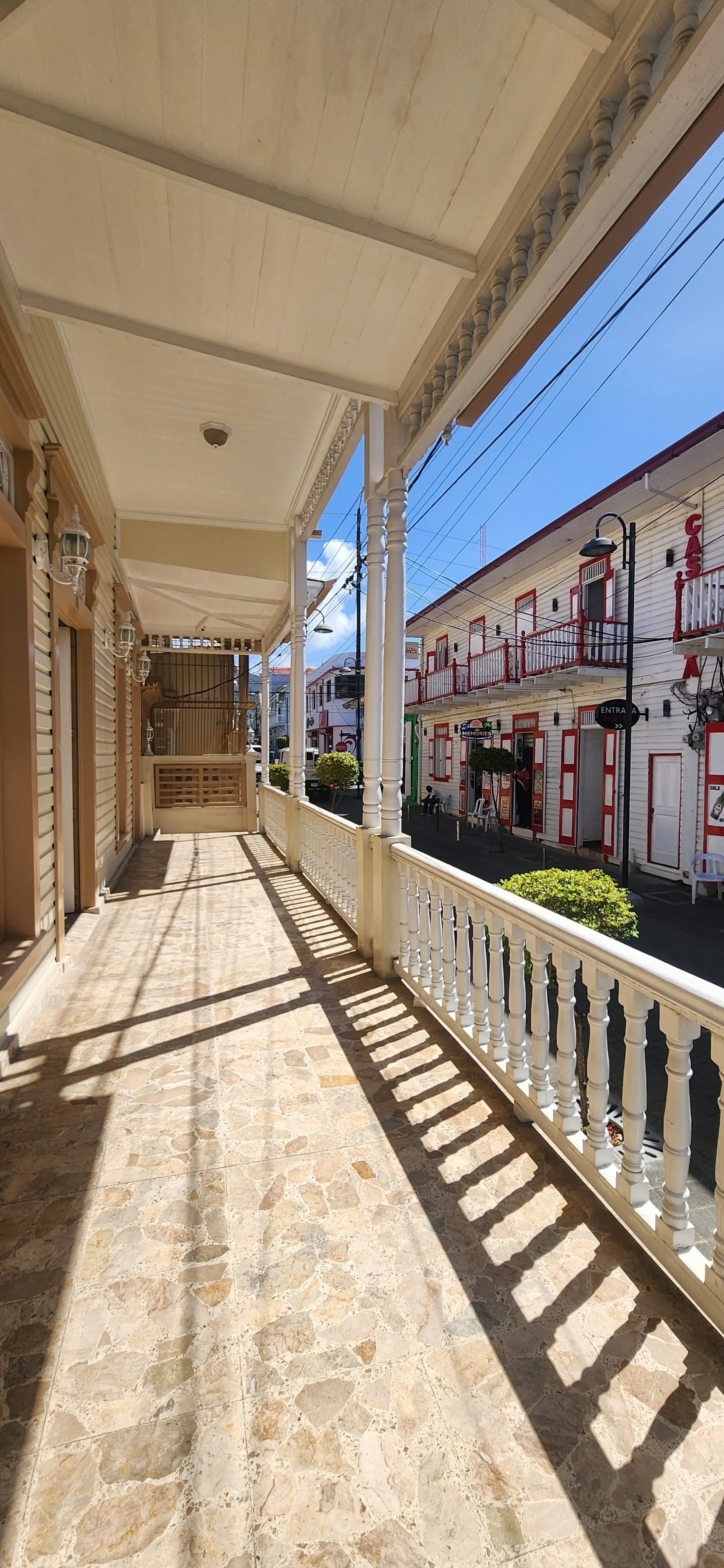 View from a balcony with beige tile flooring, white wooden railing, and a ceiling, overlooking a street with shops and buildings.