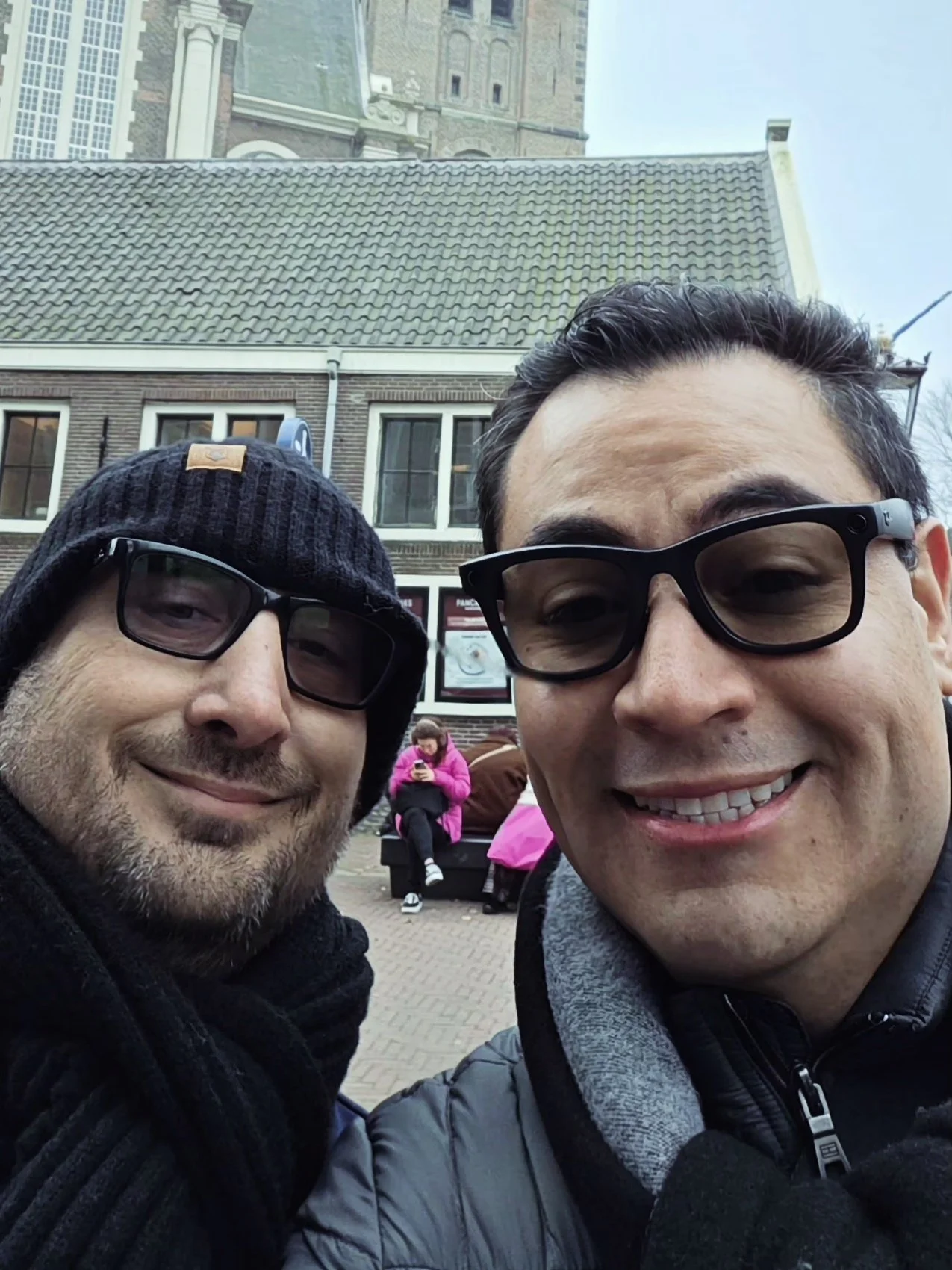 Two men smiling and wearing glasses taking a selfie outdoors in front of a historic building with a tiled roof and a tower, with a woman sitting on a bench in the background.
