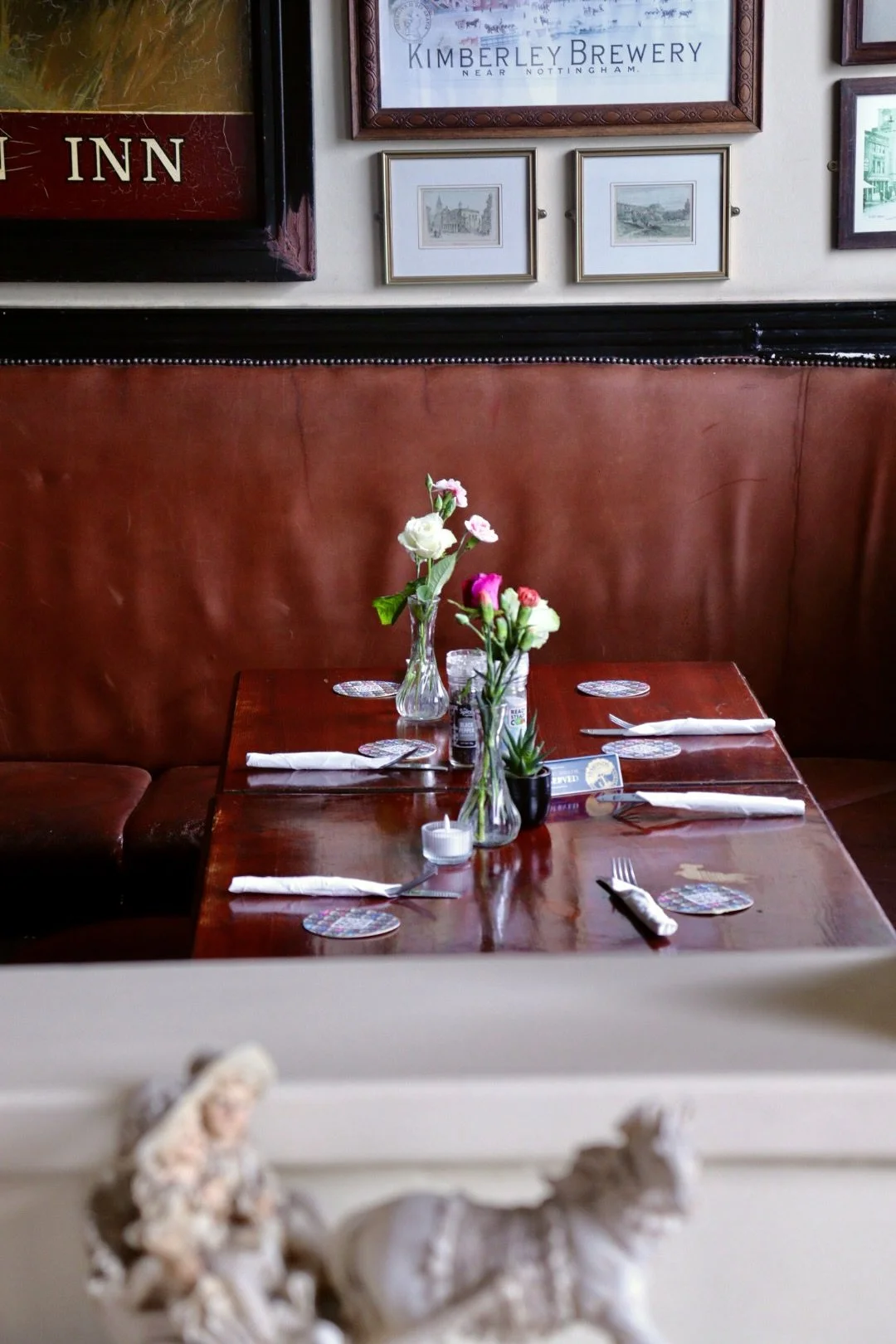 A cosy restaurant table set with vases of flowers, napkins, utensils, and coasters amid a leather booth with framed pictures and signs on the wall.