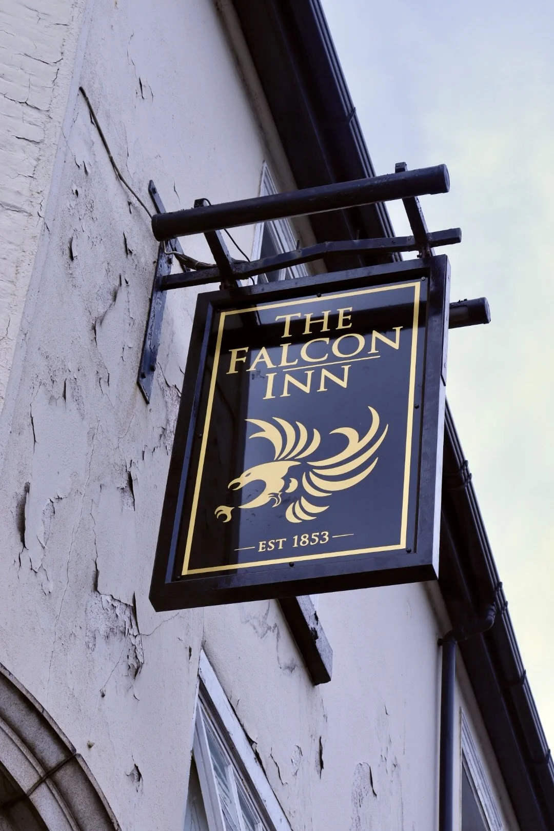 Black signboard for The Falcon Inn with gold lettering and falcon emblem, mounted on a cream-colored brick wall.
