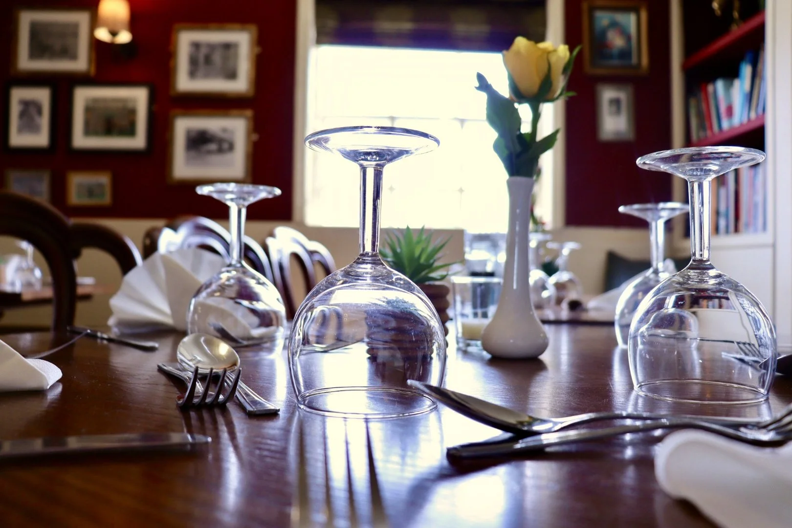 A table set for dining with upside-down wine glasses, silverware, a white napkin, and a white vase with a yellow rose. In the background, a window and framed pictures on the wall.