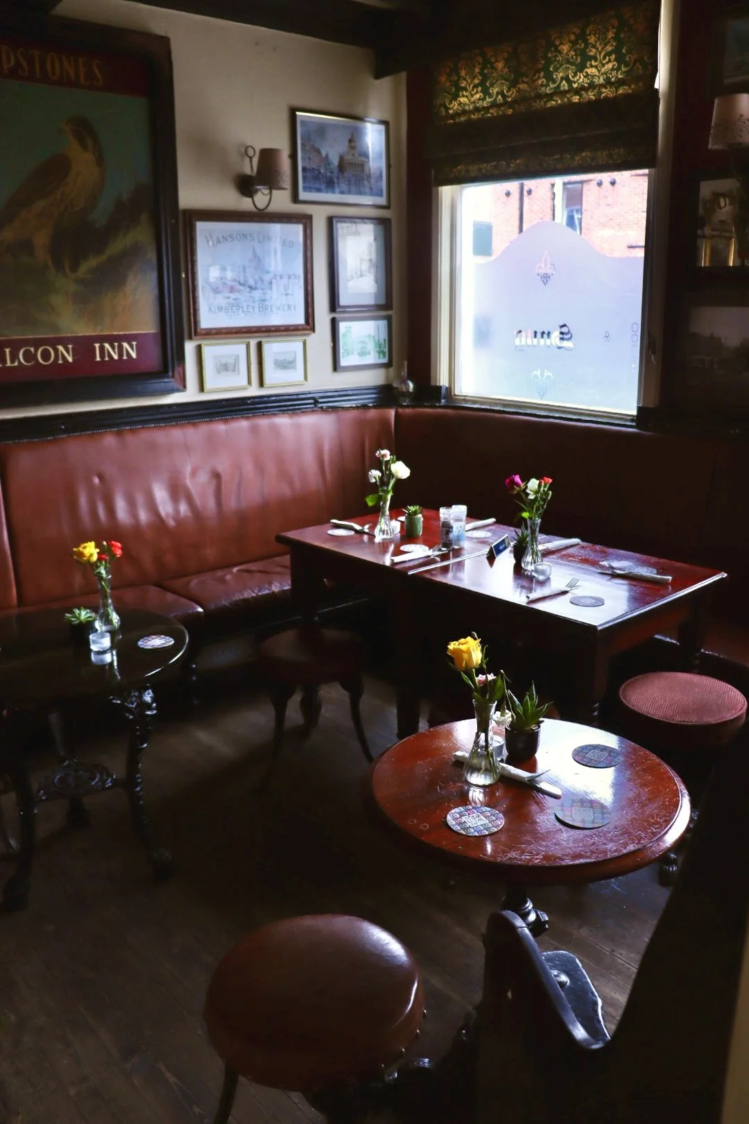 A corner with wooden tables and red leather cushioned seating. Decor includes vases with flowers on tables, framed pictures on the wall, and a window with a frosted design. The setting is warm and inviting with mixed antique and vintage decor element