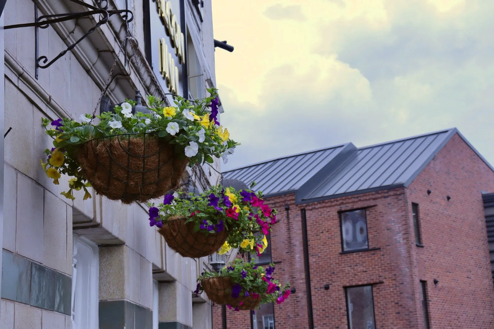Three hanging flower baskets with colorful flowers including white, yellow, purple, and pink, attached to the exterior of a building with brick and stone walls, under a cloudy sky.