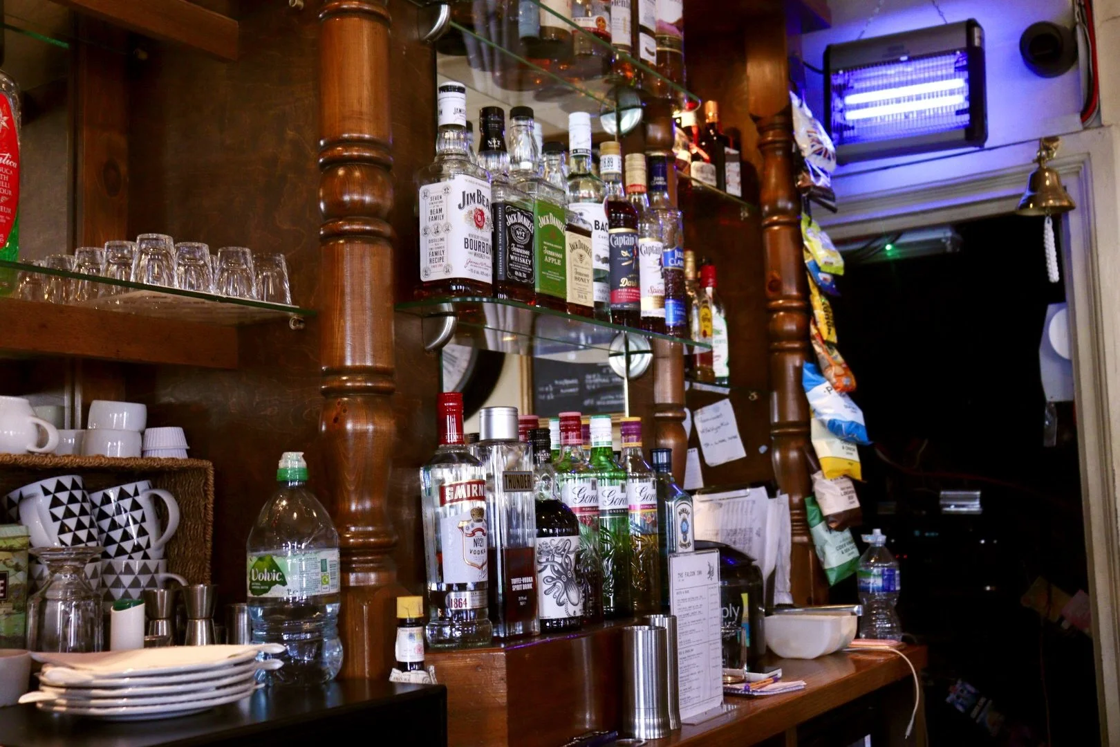 Bar shelf with various bottles of alcohol, glasses, cups, and snacks in a cosy setting.