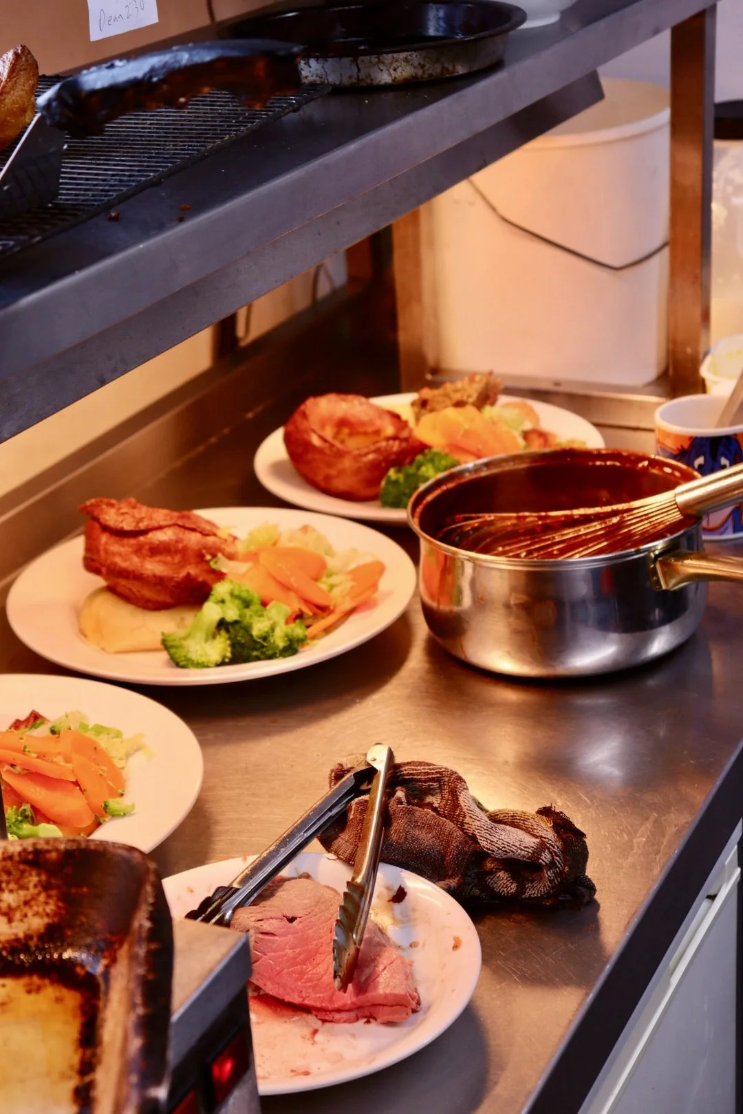 Stations with plates of roasted chicken, broccoli, carrots, and mashed potatoes, along with a pot of gravy and slices of roast beef, set on a stainless steel counter in a kitchen.