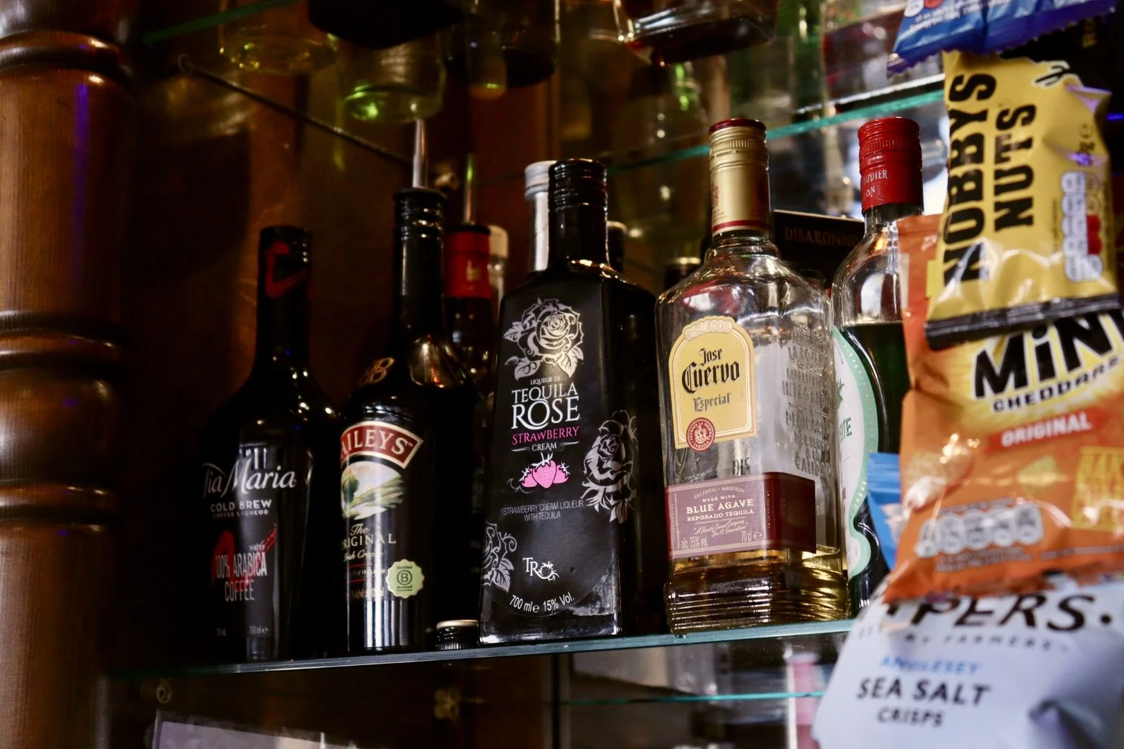 A collection of liquor bottles on a glass shelf, including tequila, liqueur, and other spirits, with a bag of sea salt chips partially visible on the right.
