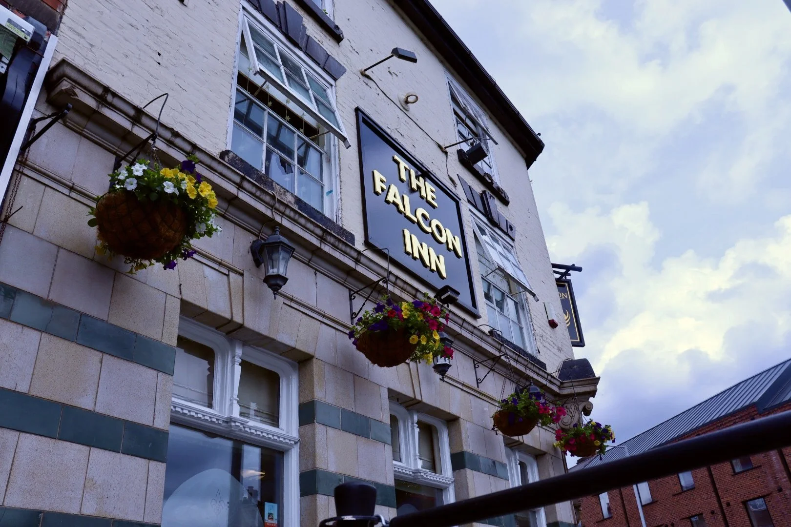 Exterior view of The Falcon Inn, a pub with hanging flower baskets and a black sign with gold lettering, under a cloudy sky.