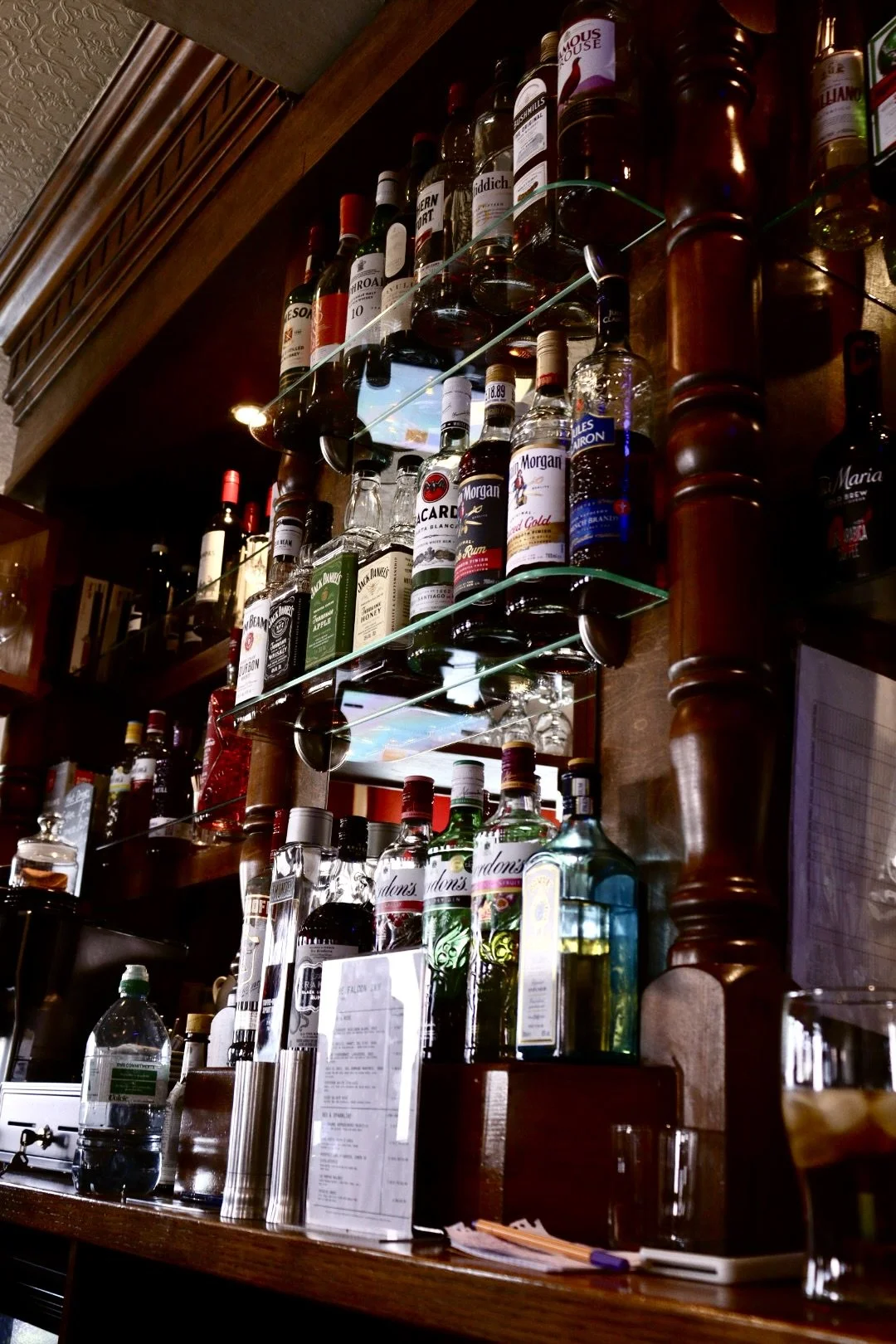A bar shelf holding various bottles of liquor, including whiskey, rum, gin, and other spirits, on glass shelves against a wooden wall.