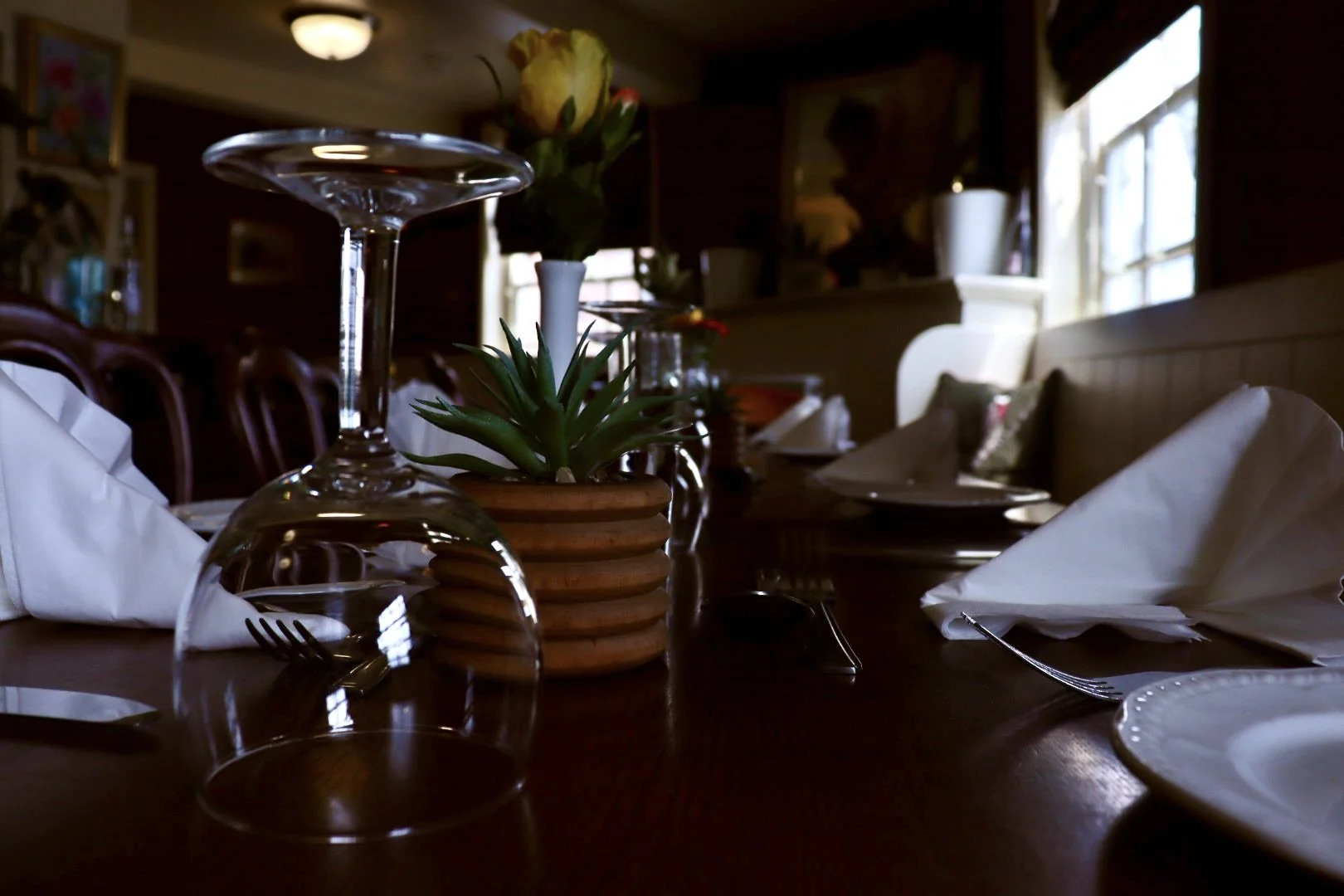A dining table set with upside-down wine glasses, folded white napkins, a small potted succulent, and a terracotta pot holding a yellow flower. In the background, there's a window letting in natural light, wooden chairs, and framed pictures on the wa