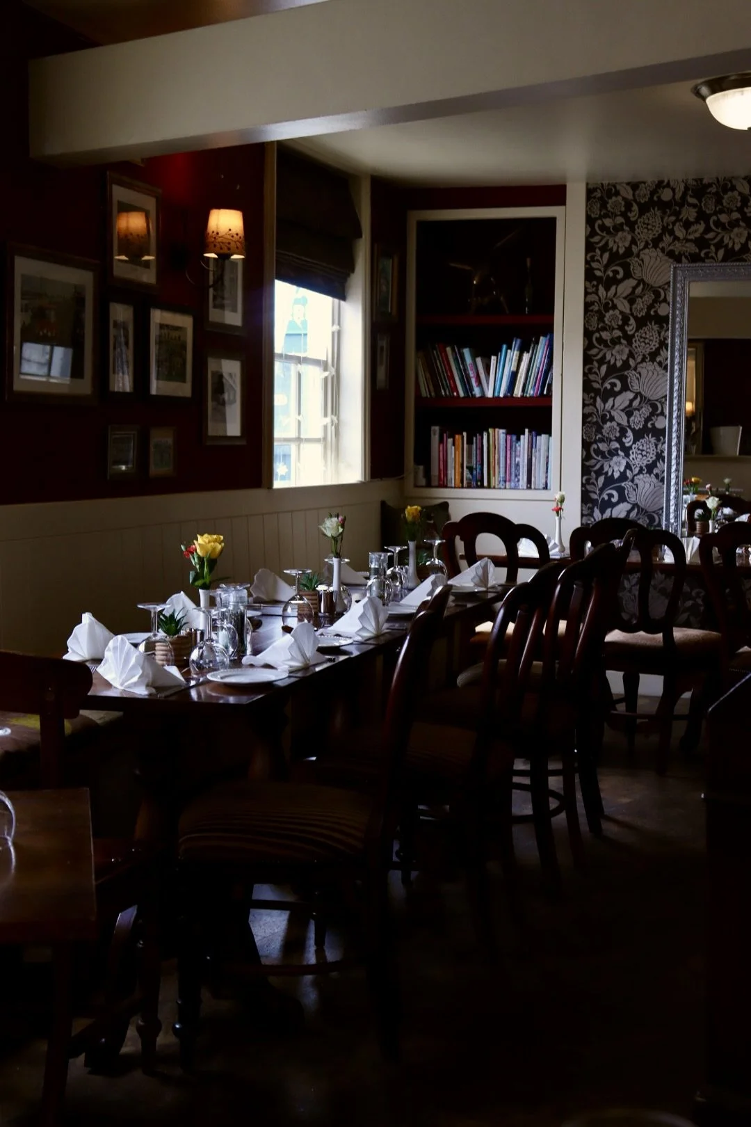 A dimly lit dining room with a long wooden table set with white napkins, glassware, and small flower arrangements. The room features a bookshelf with colorful books, a window with closed blinds, framed pictures on the wall, and decorative wallpaper w