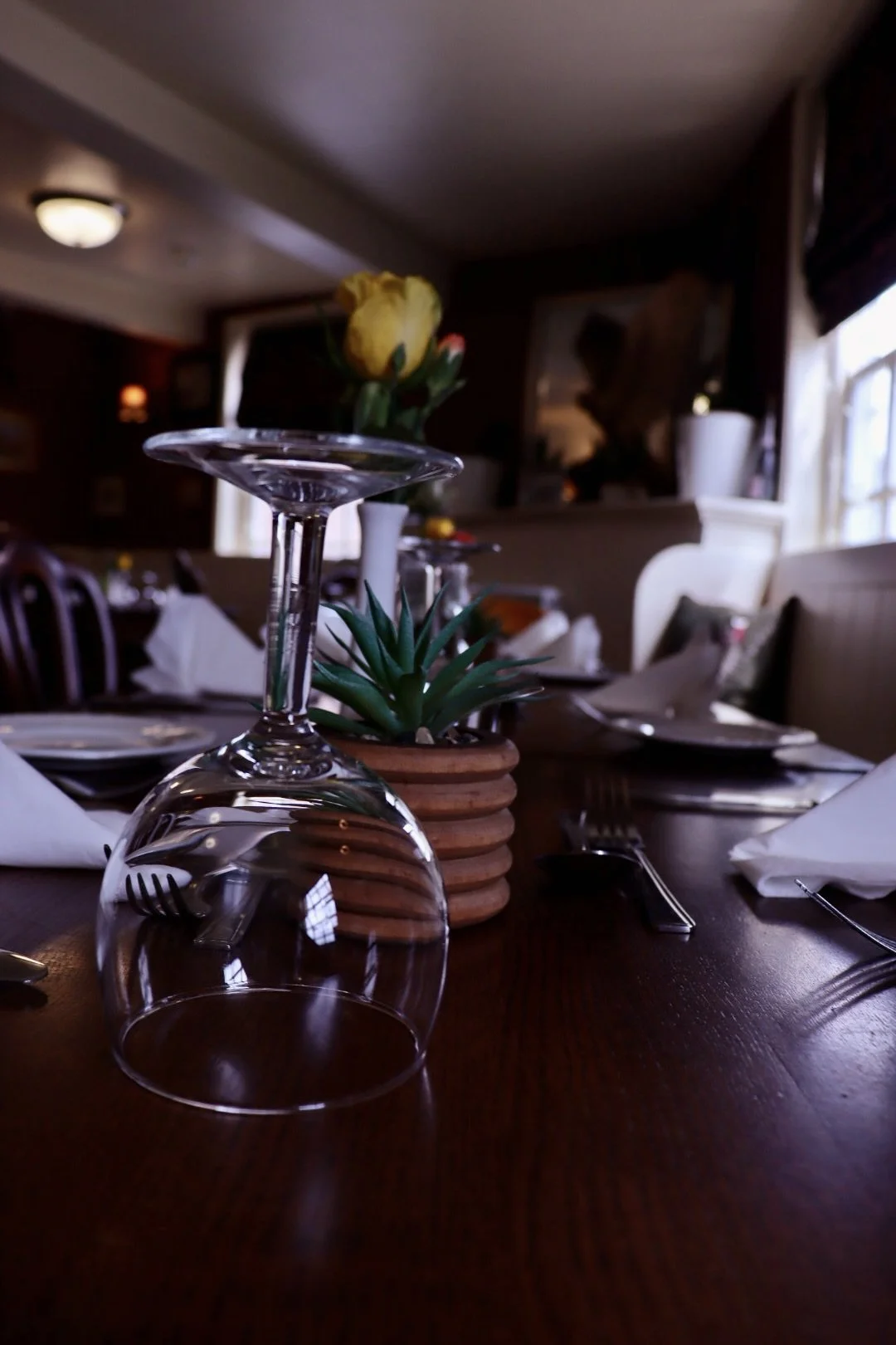 Upside-down clear wine glass on a dining table, with a small potted plant and a yellow flower in the background, set in a dimly lit restaurant with dark wood furniture and tableware.