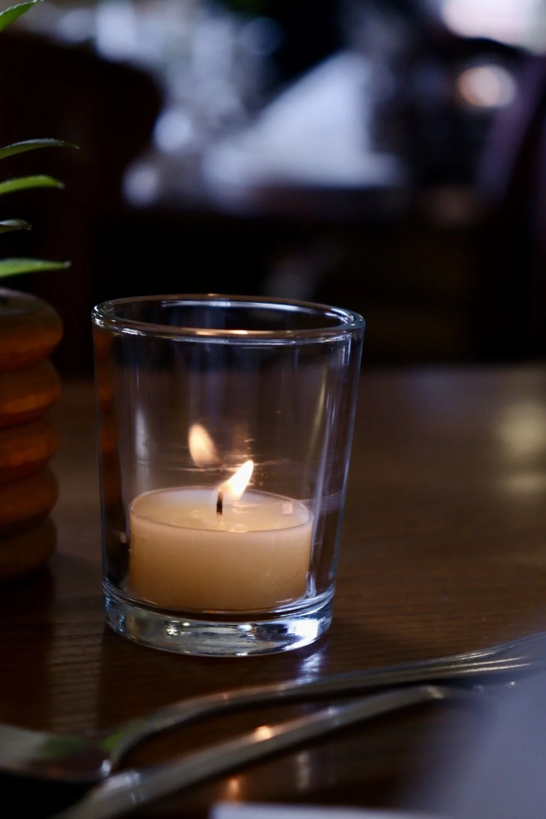 A lit candle inside a glass holder on a wooden table, with a spoon and a blurred background.